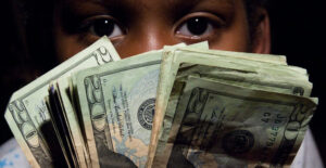 Blog The Federal Reserve creates money: Image of a young man looking at the camera from behind a "fan out" of 20 dollar bills